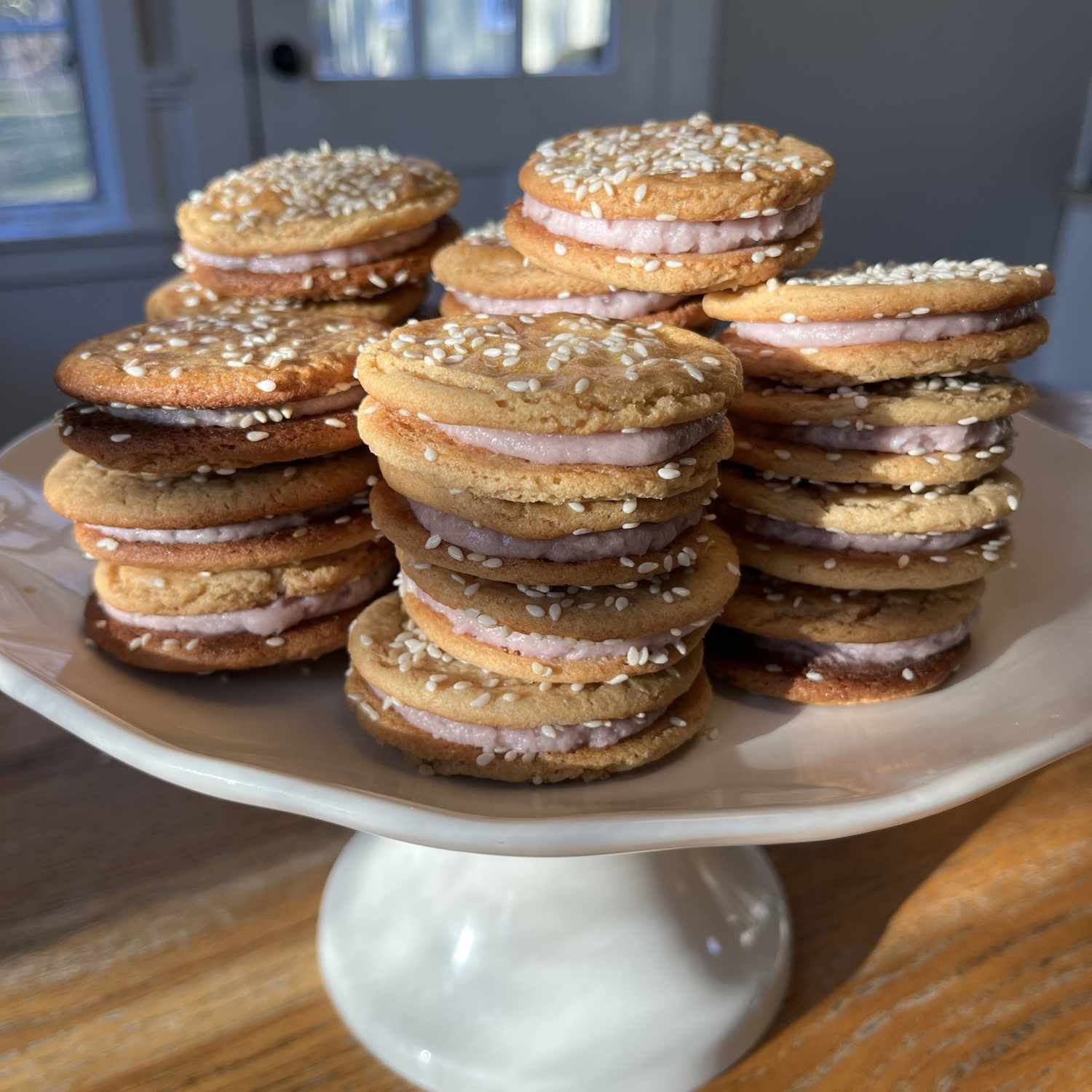 A stack of sandwich cookies garnished with sesame seeds and filled with pink filling on a white cake platter
